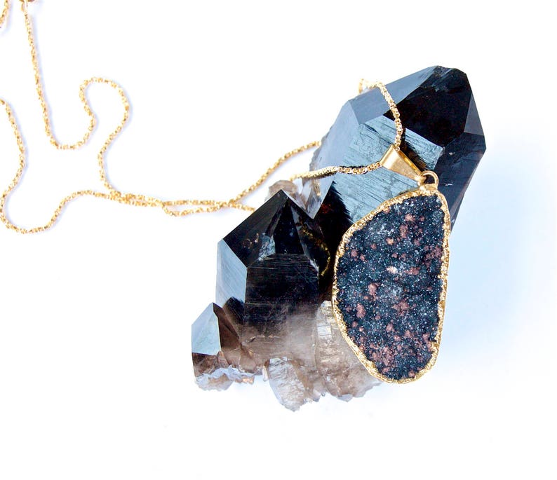 May include: A gold chain necklace with a large, dark gray, irregularly shaped pendant. The pendant is surrounded by clear and black quartz crystals.