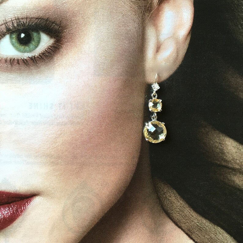 May include: A pair of silver dangle earrings with three yellow gemstones. The earrings are on a model's ear.