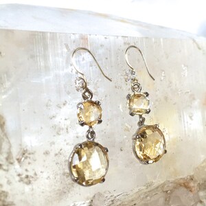 May include: A pair of silver dangle earrings with three faceted, yellow citrine stones. The stones are set in silver bezels and the earrings have a simple, elegant design.