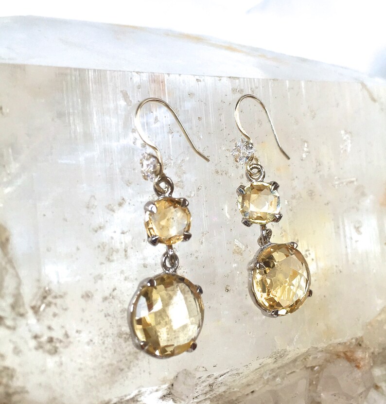 May include: A pair of silver dangle earrings with three faceted, yellow citrine stones. The stones are set in silver bezels and the earrings have a simple, elegant design.