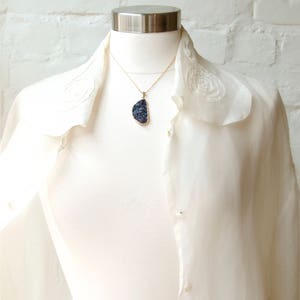 May include: A white blouse with a ruffled collar and a gold chain necklace with a large, dark blue, iridescent gemstone pendant.