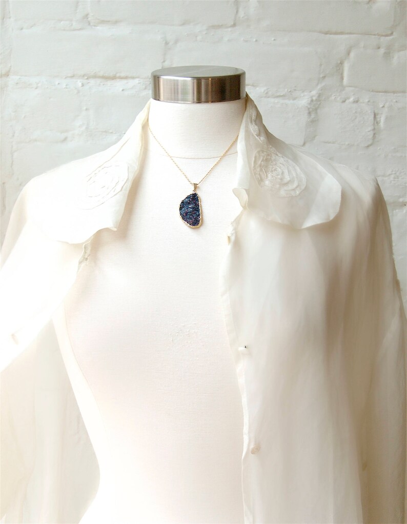 May include: A white blouse with a ruffled collar and a gold chain necklace with a large, dark blue, iridescent gemstone pendant.