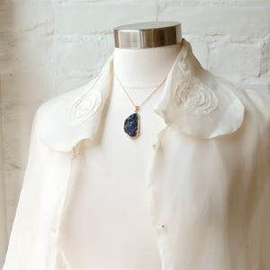 May include: A white blouse with a ruffled collar and a gold chain necklace with a dark blue stone pendant.
