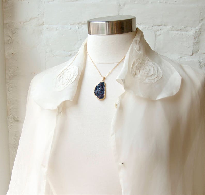 May include: A white blouse with a ruffled collar and a gold chain necklace with a dark blue stone pendant.