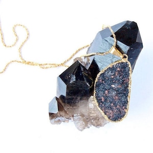 May include: A gold chain necklace with a large, dark gray, irregularly shaped pendant. The pendant is set in gold and has a speckled texture. The necklace is displayed on a cluster of black and clear quartz crystals.