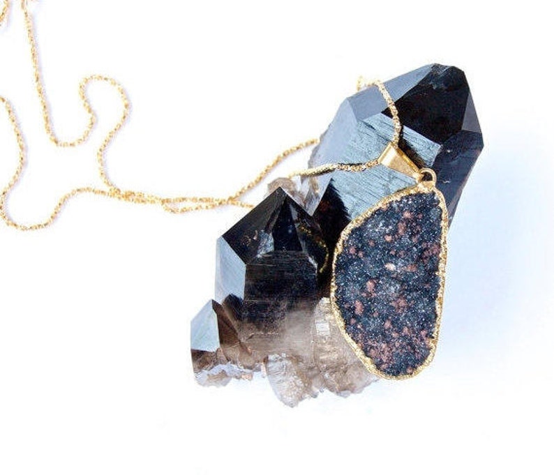 May include: A gold chain necklace with a large, dark gray, irregularly shaped pendant. The pendant is set in gold and has a speckled texture. The necklace is displayed on a cluster of black and clear quartz crystals.