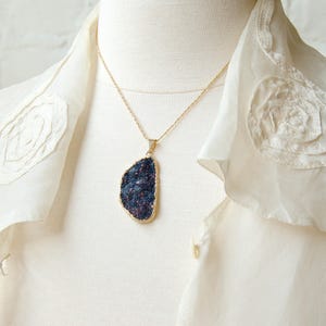 May include: A gold chain necklace with a dark blue and purple gemstone pendant. The pendant is a large, irregular shape and has a rough, textured surface. The necklace is worn on a white blouse with lace detailing.