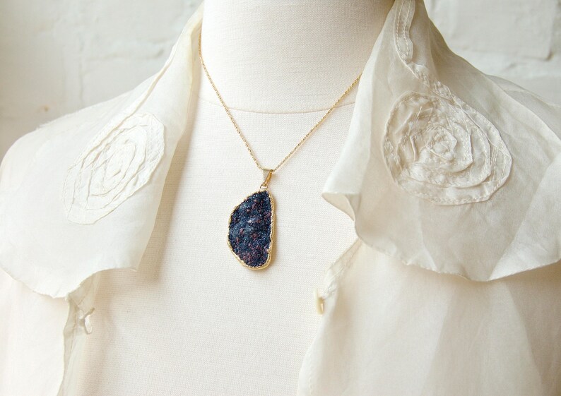 May include: A gold chain necklace with a dark blue and purple gemstone pendant. The pendant is a large, irregular shape and has a rough, textured surface. The necklace is worn on a white blouse with lace detailing.