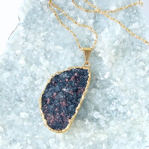 May include: A gold chain necklace with a large, dark gray and copper-colored stone pendant. The pendant is shaped like a teardrop and has a rough, textured surface. The necklace is displayed on a bed of white crystals.