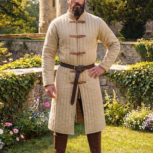May include: A beige, knee-length quilted gambeson featuring vertical stitching, a high collar, and a front closure secured by three brown leather straps with metal buckles. A dark brown leather belt is fastened around the waist over the garment.