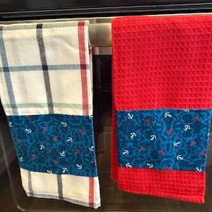 May include: Two kitchen towels hanging on a silver oven rack. The towel on the left is white with black and red plaid and a blue and white anchor print. The towel on the right is red with a blue and white anchor print.