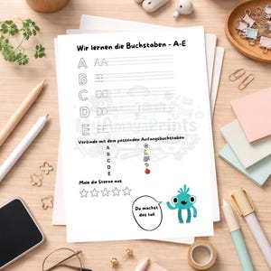 May include: A white worksheet with the text "Wir lernen die Buchstaben - A-E" and letter tracing exercises. The worksheet includes illustrations and the phrase "Du machst das toll". The image is on a wooden surface with stationery.