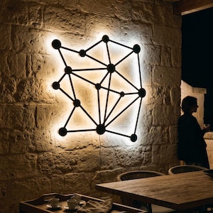 May include: A wall-mounted geometric light fixture featuring a black frame with interconnected lines and circular nodes, emitting a warm white glow against a textured stone wall. The design resembles a network or molecular structure.