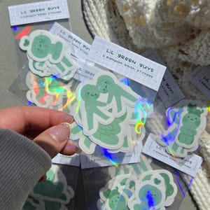 May include: Multiple packs of stickers with light green cartoon figures. Each pack is labelled "LIL GREEN GUYS" and "5 RANDOM smiski STICKERS". The stickers have a holographic finish.