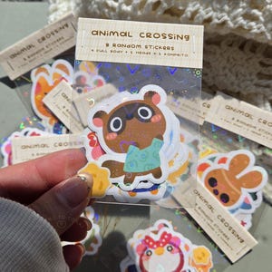 May include: A pack of Animal Crossing stickers in a clear plastic sleeve. The stickers showcase characters from the game with a holographic finish. The label on the pack states "Animal Crossing" and "8 Random Stickers."