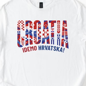 May include: White t-shirt with the word "CROATIA" in red and blue decorative lettering. Below the word is the phrase "IDEMO HRVATSKA!" in blue. The shirt is a crew neck style.