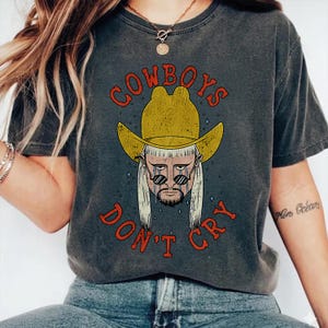 May include: A charcoal gray t-shirt featuring a graphic print of a person with a mullet hairstyle wearing a yellow cowboy hat and sunglasses. The text "COWBOYS DON'T CRY" is printed in red above and below the illustration, with blue teardrops.