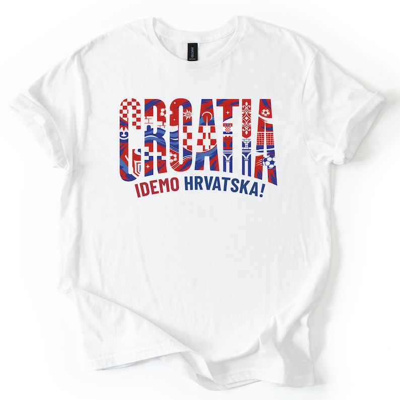 May include: White t-shirt with the word "CROATIA" in red and blue decorative lettering. Below the word is the phrase "IDEMO HRVATSKA!" in blue. The shirt is a crew neck style.
