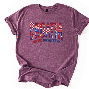 Puede incluir: Una camiseta granate con la palabra "CROATIA" en rojo y azul, dise&ntilde;ada con varios s&iacute;mbolos croatas. Debajo est&aacute; la frase "IDEMO HRVATSKA!" en azul. La camiseta est&aacute; hecha de un material suave.