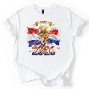 Może przedstawiać: Biała koszulka z nadrukiem złotego trofeum, chorwackiej flagi i napisem "Croatia 2026". Projekt zawiera złote akcenty i wstążkę z napisem "Croatia".