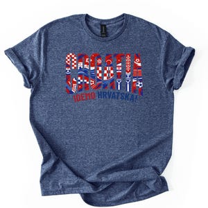 Puede incluir: Una camiseta azul marino con un dise&ntilde;o gr&aacute;fico que presenta la palabra "CROACIA" en rojo, blanco y azul, junto con varios s&iacute;mbolos croatas y el texto "IDEMO HRVATSKA!". La camiseta tiene mangas enrolladas y cuello redondo.