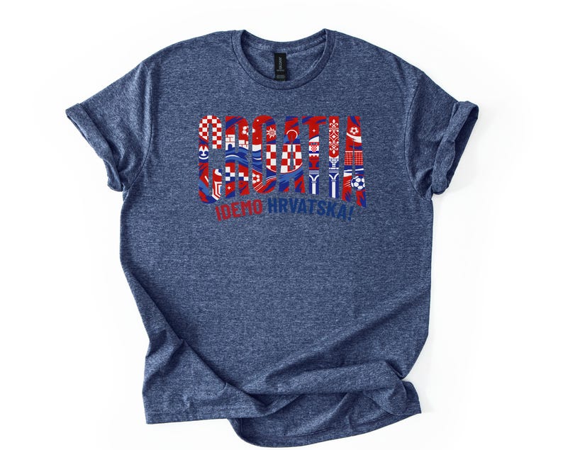 Puede incluir: Una camiseta azul marino con un dise&ntilde;o gr&aacute;fico que presenta la palabra "CROACIA" en rojo, blanco y azul, junto con varios s&iacute;mbolos croatas y el texto "IDEMO HRVATSKA!". La camiseta tiene mangas enrolladas y cuello redondo.