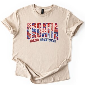 Puede incluir: Camiseta beige con la palabra "CROATIA" en rojo y azul, estilizada con patrones de la bandera croata. Debajo, el texto "IDEMO HRVATSKA!" en rojo. La camiseta es de material suave.