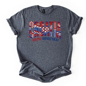 Puede incluir: Una camiseta gris jaspeada con la palabra "SOCCER" en rojo, blanco y azul, con dise&ntilde;os inspirados en la bandera croata. Debajo de la palabra "SOCCER" est&aacute; el texto "IDEMO HRVATSKA!" en rojo. La camiseta es de material suave.
