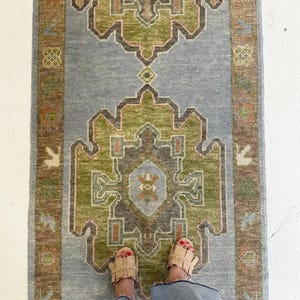 Könnte beinhalten: Ein rechteckiger Teppich mit einem zentralen Design in Grün-, Braun- und Orangetönen auf hellblauem Hintergrund. Der Teppich hat ein kompliziertes, geometrisches Muster und einen braunen Rand. Draufsicht.