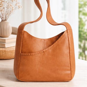 May include: A tan-colored handbag with a curved handle. The bag has a rectangular shape with a curved top and a front pocket. The bag is made of a smooth material and sits on a wooden table. The bag is a neutral color and could be used for everyday use.