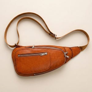 Puede incluir: Un bolso de cintura de cuero marrón con múltiples compartimentos con cremallera y una correa ajustable de color beige. El bolso tiene un bolsillo frontal rectangular y una forma curva. Las cremalleras son plateadas.