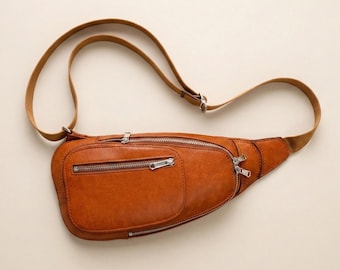 Puede incluir: Un bolso de cintura de cuero marrón con múltiples compartimentos con cremallera y una correa ajustable de color beige. El bolso tiene un bolsillo frontal rectangular y una forma curva. Las cremalleras son plateadas.