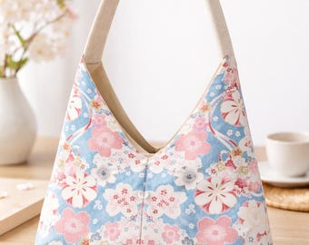 Puede incluir: Bolso con estampado floral con flores rosas y blancas sobre un fondo azul claro. Tiene un asa beige y una forma triangular distintiva. El bolso está sobre una superficie de madera.