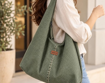 Puede incluir: Un bolso de hombro estilo hobo en color verde salvia confeccionado en tela texturizada. El bolso cuenta con una correa ancha, una costura vertical central y un pequeño parche rectangular de aspecto cuero marrón en el frente.