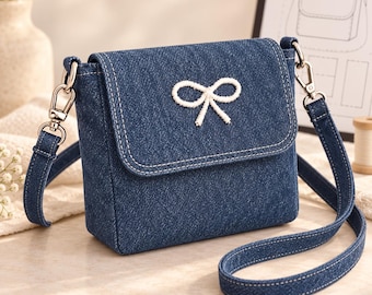 Puede incluir: Un bolso bandolera de mezclilla azul oscuro con una solapa rectangular, costuras blancas en contraste y un detalle de lazo bordado en blanco. El bolso incluye una correa de mezclilla ajustable a juego con cierres giratorios de metal plateado.
