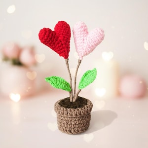 Puede incluir: Un arreglo floral de ganchillo con dos flores en forma de corazón, una roja y otra rosa. Los tallos son marrones, con hojas verdes, y la planta está en una maceta de ganchillo marrón. El fondo es borroso.