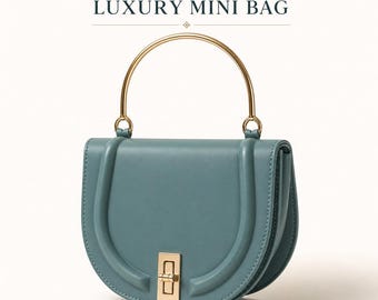 Puede incluir: Un mini bolso semicircular de color turquesa con asa y cierre dorados. El bolso está hecho de cuero liso y presenta el texto "Handmade LUXURY MINI BAG" en letras doradas.
