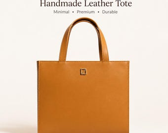 Puede incluir: Un bolso tote de cuero color marrón claro con asas cortas. El bolso tiene forma rectangular con un pequeño emblema metálico cuadrado en la parte delantera. El texto "Handmade Leather Tote" está encima del bolso, con las palabras "Minimal, Premium, Durable" debajo.