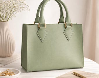 Puede incluir: Un bolso de mano estructurado en color verde salvia con dos asas superiores, que presenta una silueta rectangular y herrajes en tono dorado en los puntos donde las asas se unen al cuerpo principal del bolso, creando un diseño limpio.