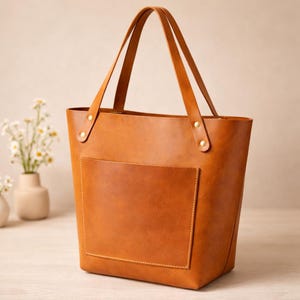 May include: A tan leather tote bag with a front pocket and two handles. The bag has a rectangular shape and is secured with rivets at the top. The bag is made of a smooth, solid-coloured leather.