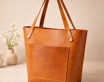 Puede incluir: Un bolso tote de cuero marrón con un bolsillo frontal y dos asas. El bolso tiene forma rectangular y está asegurado con remaches en la parte superior. El bolso está hecho de cuero liso de color sólido.