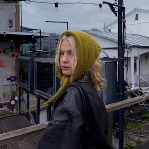 Könnte beinhalten: Eine gestrickte olivgrüne Kapuze mit weicher, strukturierter Oberfläche, die den Kopf bedeckt und unter dem Kinn gebunden wird. Die Kapuze wird über einer schwarzen Lederjacke getragen und zeichnet sich durch eine einfache Strickstruktur aus.