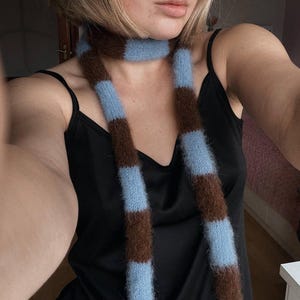 Puede incluir: Una bufanda larga a rayas con bandas alternas de azul claro y marrón. La bufanda está hecha de un material suave y difuso y se lleva alrededor del cuello. La bufanda es un accesorio de moda.