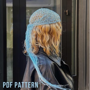 May include: A light blue crochet headscarf featuring an open-knit mesh pattern and long, dangling fringe strands that extend down the side. The headscarf is worn over wavy blonde hair and is accompanied by the text PDF PATTERN at the bottom.
