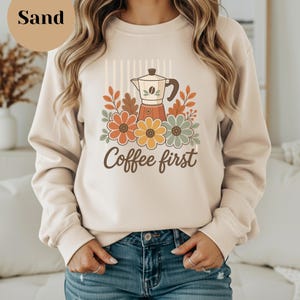 May include: A sand-colored crewneck sweatshirt featuring a graphic print of a moka pot surrounded by orange, yellow, and sage green flowers and leaves. The text Coffee first is printed in brown script below the floral design on the front.