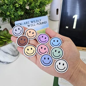 Peut inclure: Plusieurs autocollants ronds avec des visages souriants de différentes couleurs, dont le bleu, le rose et le jaune. Chaque autocollant a un visage souriant noir et un bord blanc. Le texte "YOU ARE WILDLY WELL-BOUND!" est visible en arrière-plan.