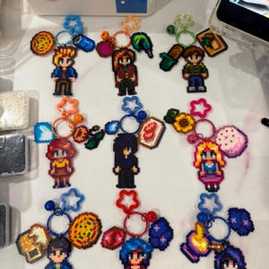 Könnte beinhalten: Eine Sammlung von Pixel-Art-Charakter-Schlüsselanhängern. Jeder Schlüsselanhänger zeigt ein anderes Charakterdesign mit verschiedenen Accessoires und Anhängern. Die Schlüsselanhänger sind an Metallringen mit bunten Stern- und Blumenakzenten befestigt. Die Charaktere tragen verschiedene Outfits.
