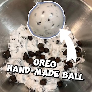 May include: A close-up shot of a white, hand-made Oreo ball with dark chocolate cookie pieces. The ball is in a stainless steel bowl, surrounded by a white mixture with dark cookie pieces. The text "OREO HAND-MADE BALL" is displayed.