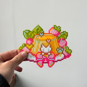 Può includere: Personaggio in pixel art con un vestito giallo, scarpe rosa e una corona di fragole e foglie. Il personaggio ha un viso bianco ed è circondato da fragole rosa e foglie verdi. L'arte è fatta di piccoli quadrati colorati.