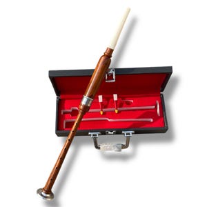 Puede incluir: Una gaita de madera con un acento plateado y una punta blanca, colocada en un estuche negro abierto con interior rojo. El estuche contiene las lengüetas y otros componentes de la gaita. Instrumento musical tradicional.